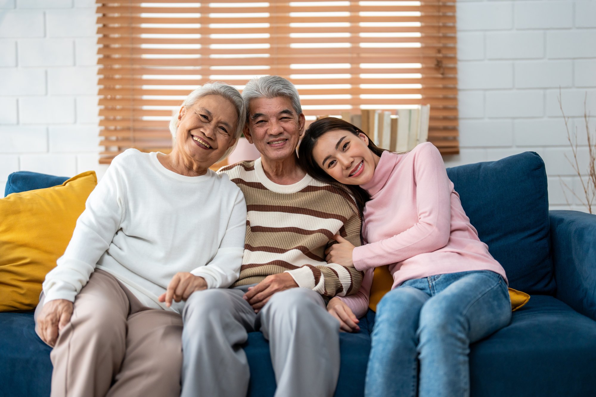 Ein glückliches älteres asiatisches Paar sitzt auf einem Sofa und wird von einer jüngeren liebevoll Frau umarmt. Die Szene strahlt Wärme, Nähe und familiäre Verbundenheit aus